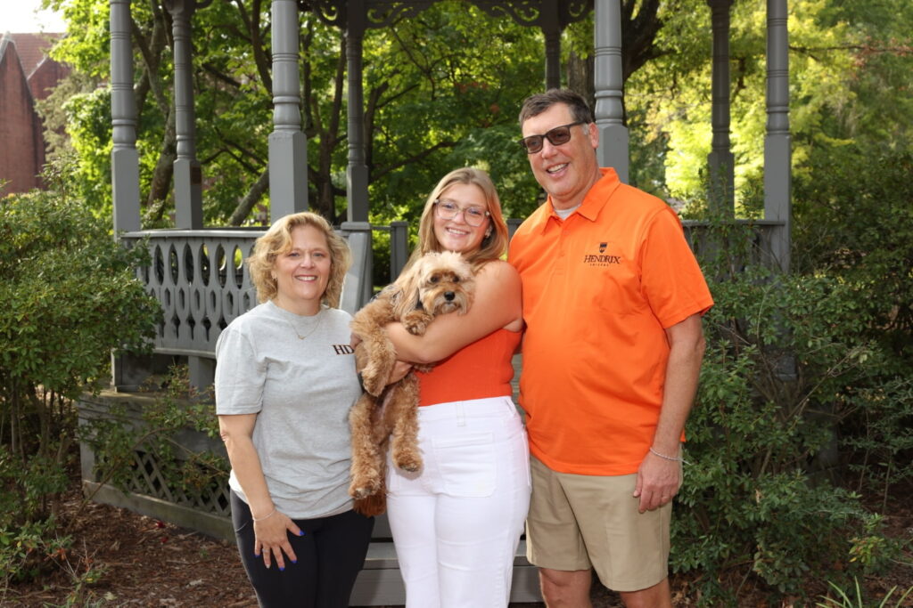 Three people smiling at the camera while standing outside in front of a gazebo. One person is holding a dog.