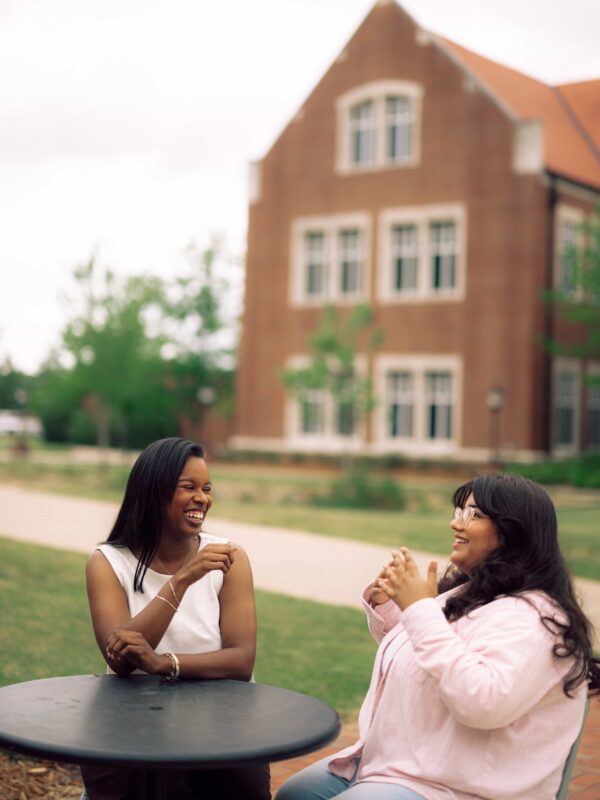 Two smiling students having a conversation at an outdoor table on campus.