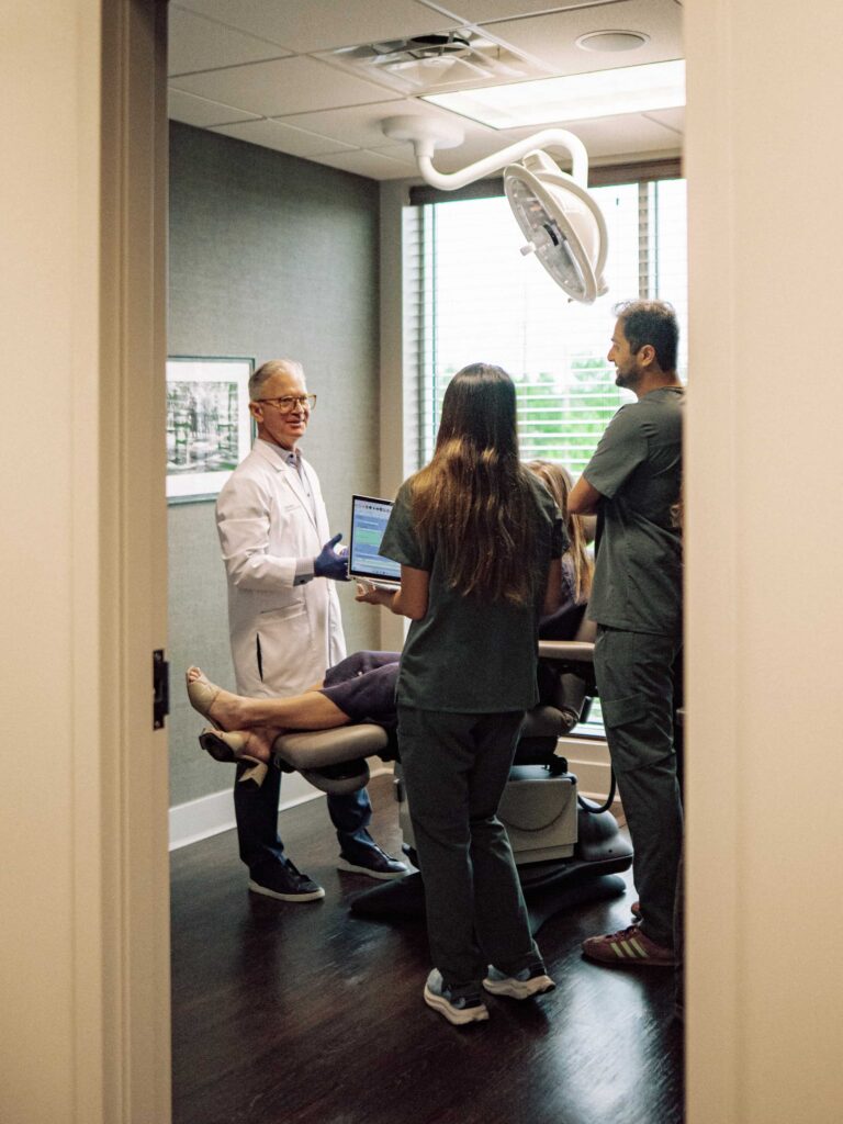 A doctor stands alongside a patient who is sitting in an exam chair while two students observe.