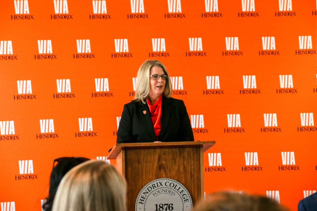 President Karen Petersen speaks at a podium during the launch of Via Hendrix. She stands in front of a large orange backdrop with the Via Hendrix logo shown.