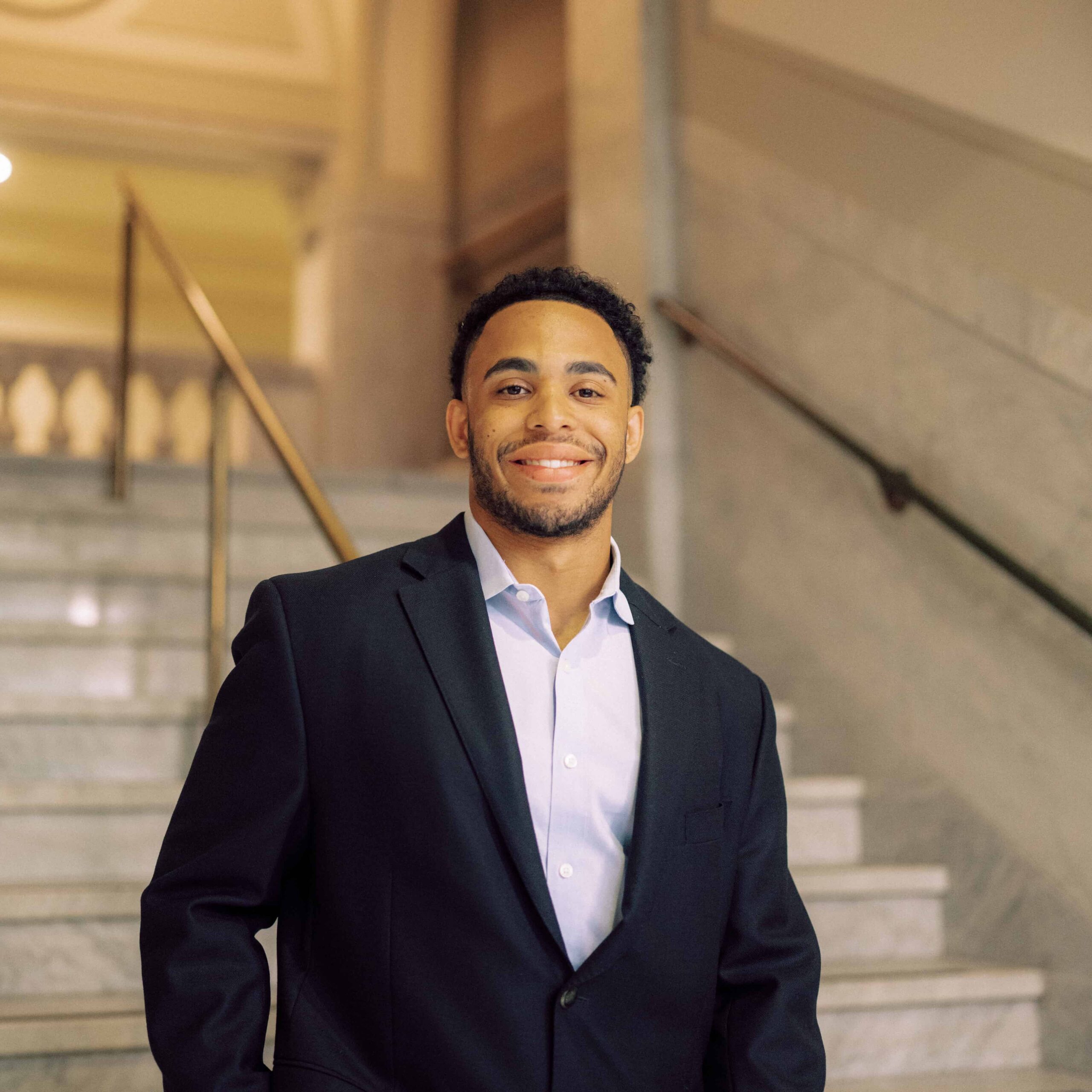 A student dressed in a business suit stands on a staircase inside a courthouse while smiling.