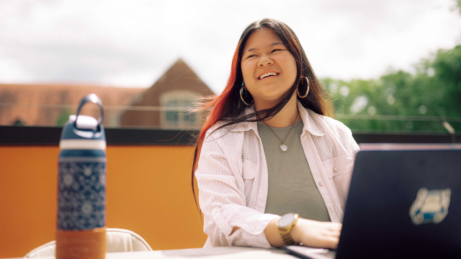 Smiling Hendrix psychology student studies outdoors at a table with a laptop and water bottle on campus.