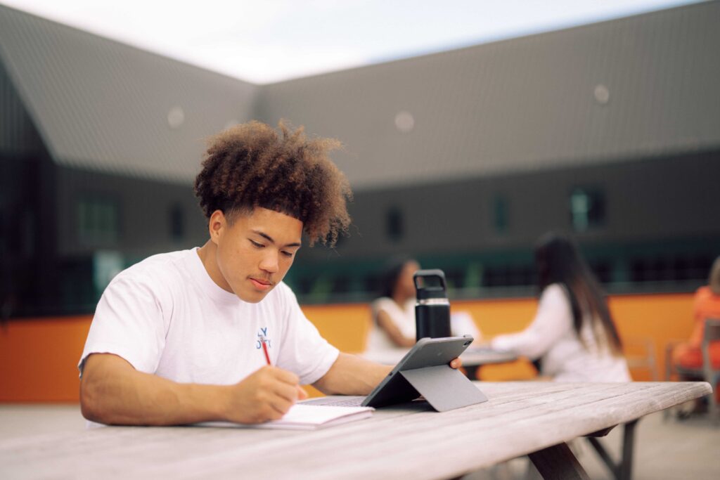 A student sitting at an outdoor table, writing in their notebook with a tablet and water bottle next to them.