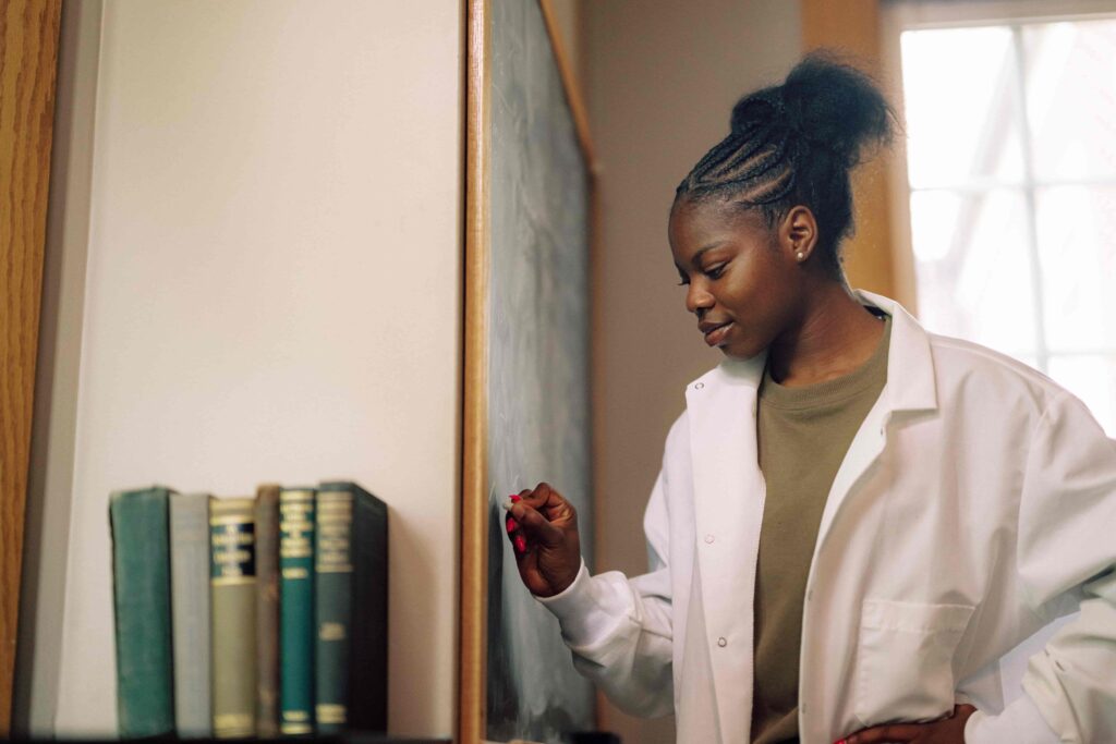 A student in a white coat writing equations on a chalkboard.