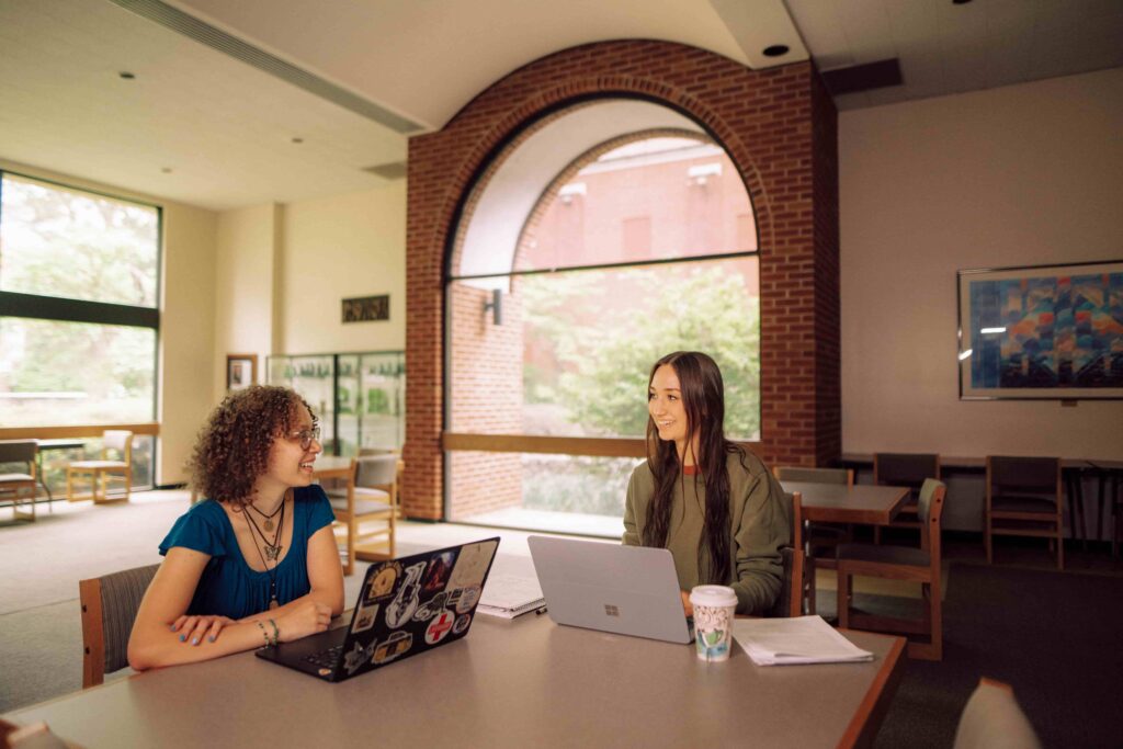 Two students studying at a table with their laptops and notes.