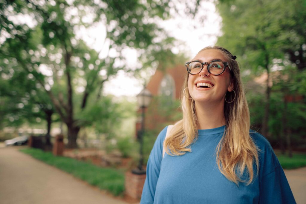 A smiling student looking up at the sky while standing on a tree lined sidewalk.