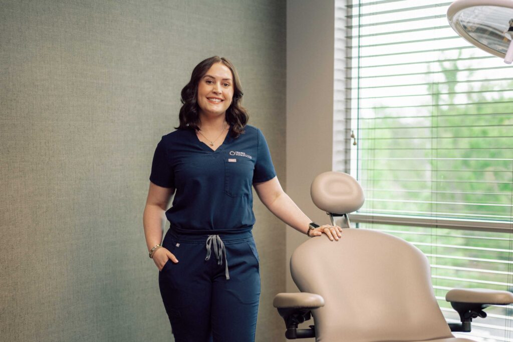 A smiling student wearing medical scrubs, standing next to a chair in a patient exam room.