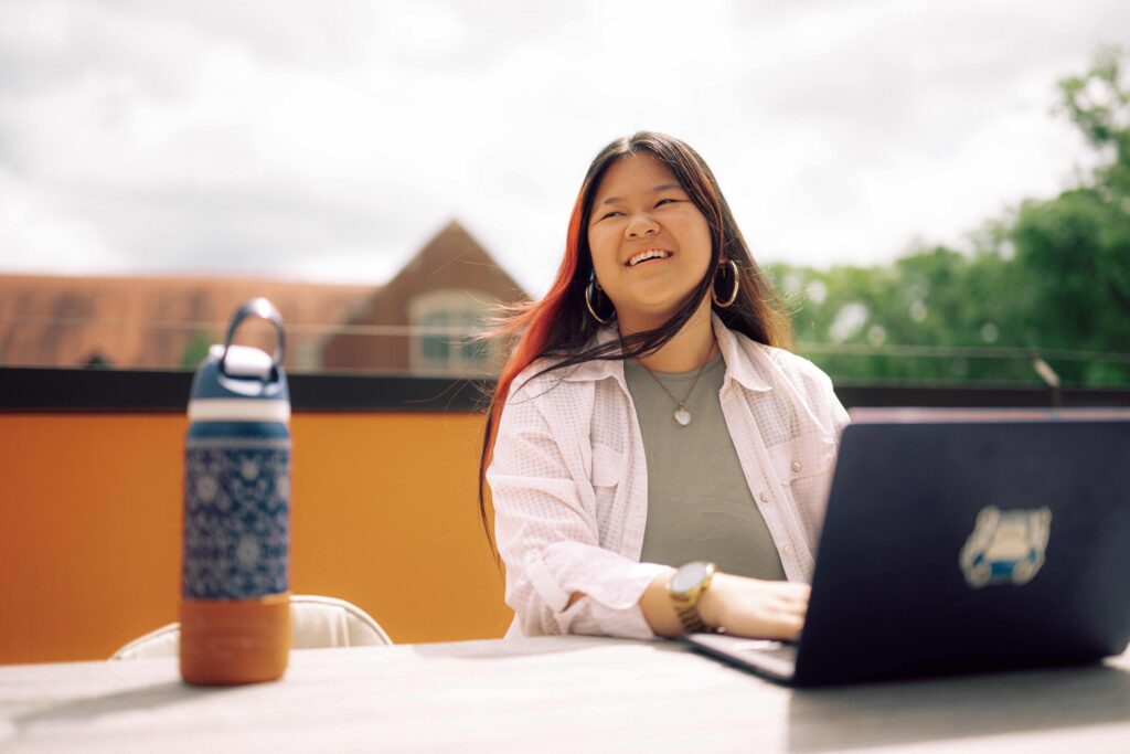 A smiling student using a laptop computer while sitting at an outdoor table.