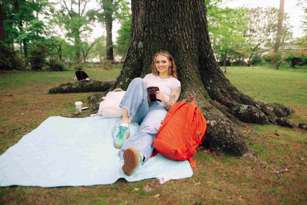 A smiling student sitting on a blanket while leaned against a tree with a book in their hands.