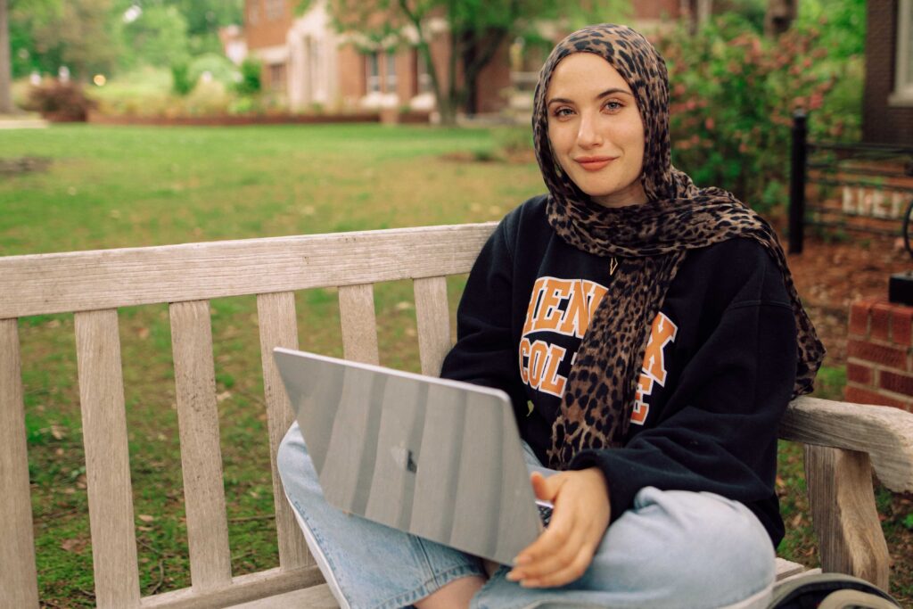A student wearing a headscarf, sitting on a wooden outdoor bench with a laptop computer in her lap.