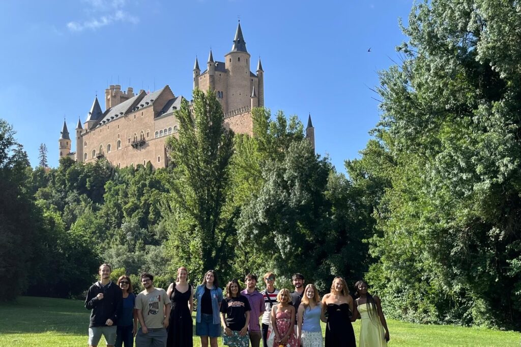 A group of thirteen students standing shoulder to shoulder with a large Spanish style castle in the background.