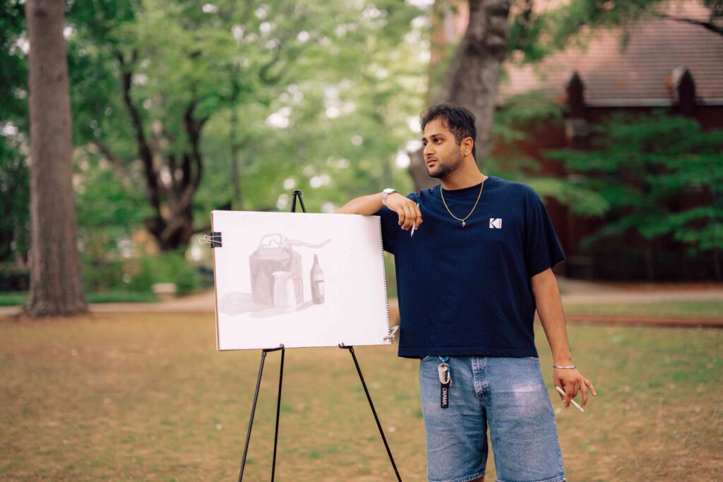 A student standing outdoors, leaning on an easel with his art on it.