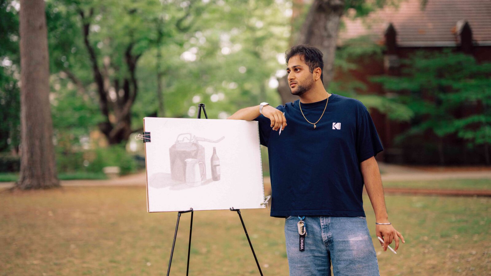 A student standing outdoors, leaning on an easel with his art on it.