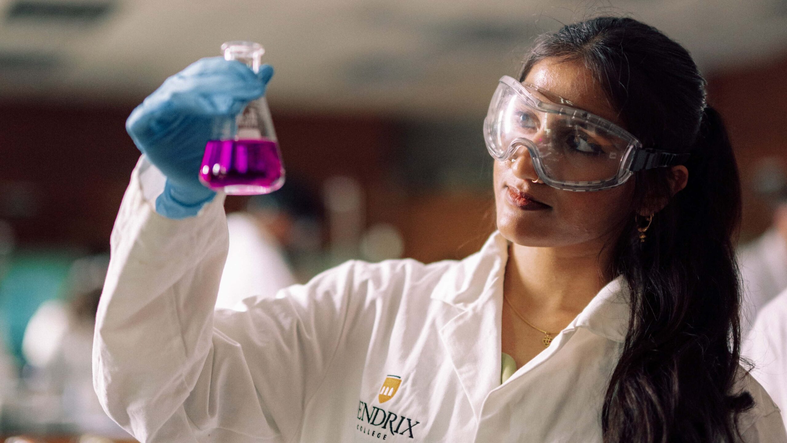 A student wearing safety equipment examining the contents of a beaker in a science lab.