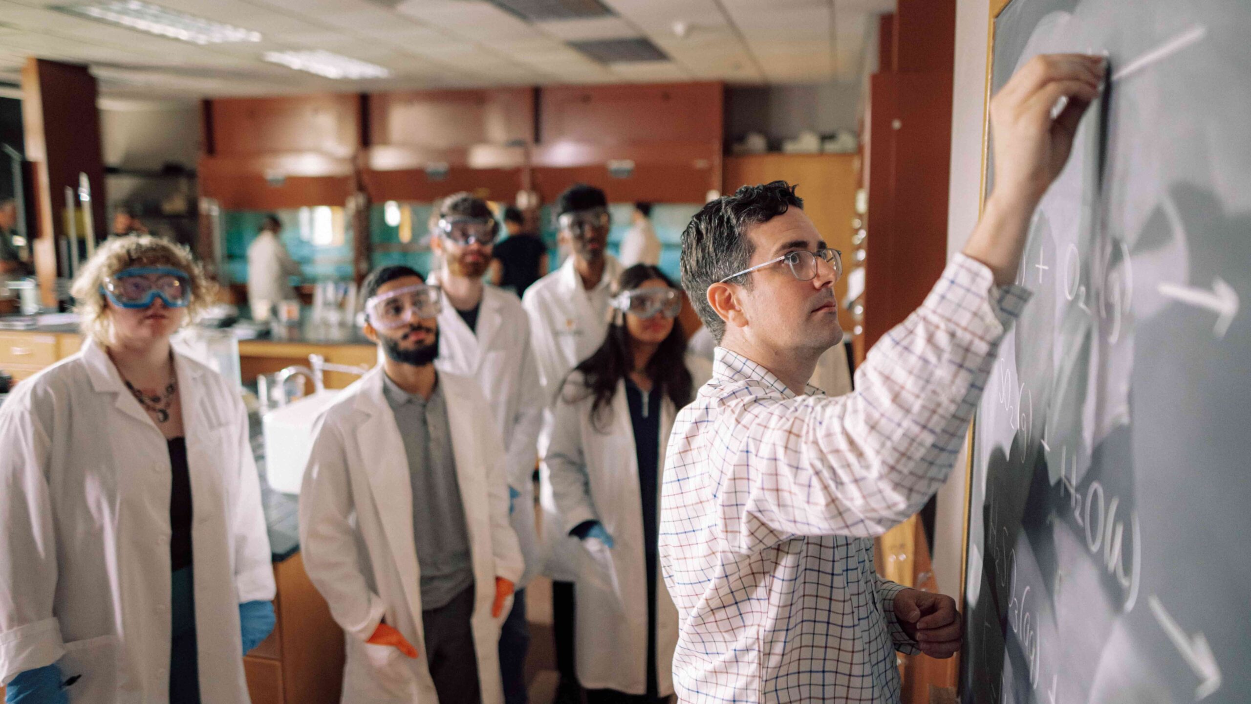 A Hendrix College professor writes equations on a chalkboard while a group of lab students in safety goggles and coats observe.