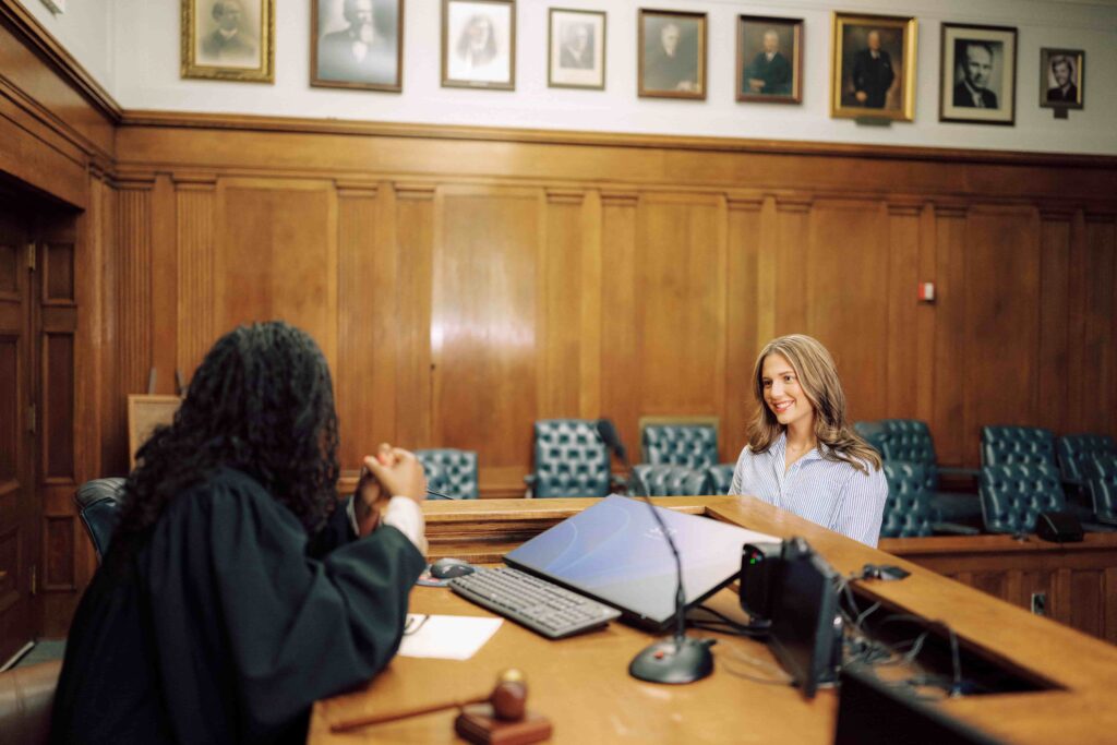 Two people talk with one another in a courtroom. One wears a black gown behind the courtroom bench.