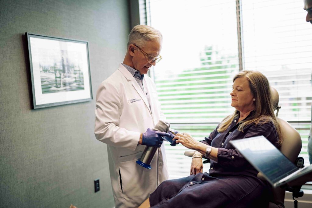 One medical professional holds a device up to another person’s finger. The other person sits in a medical examination chair holding up their hand.