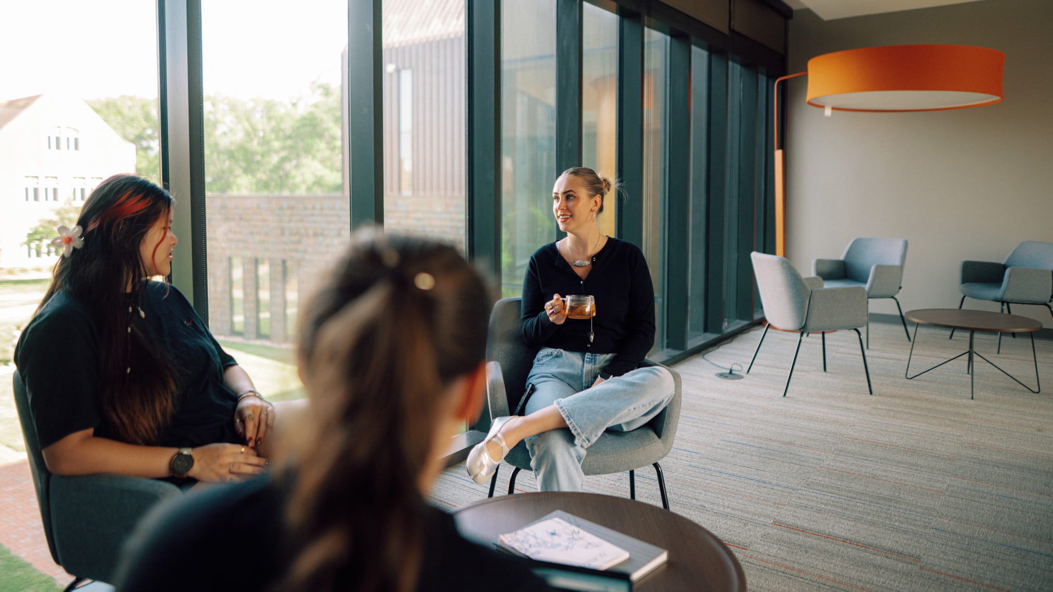 Three students sitting indoors by a glass-paneled wall with a coffee table in front of them.