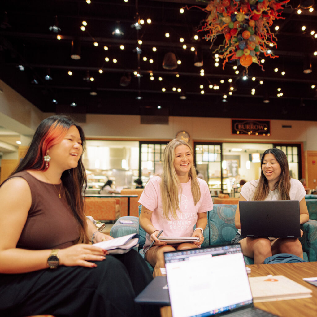 Three people sitting in a well-lit room chatting with one another around a coffee table. Two of the people have laptops in front of them.