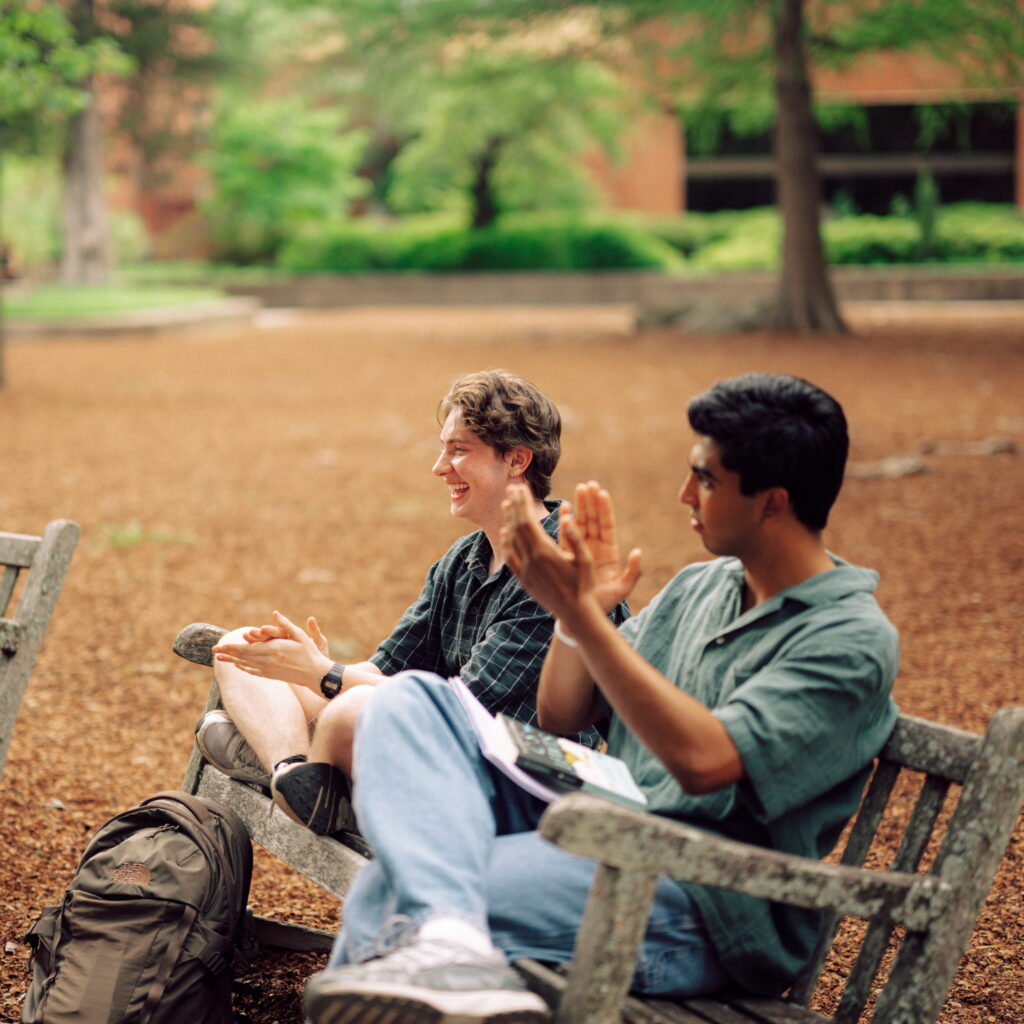 Two people sit on a wooden bench outside on a sunny day. One person is clapping.