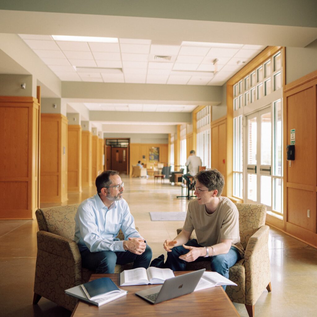 Two people sit behind a coffee table chatting with one another in a well-lit room.
