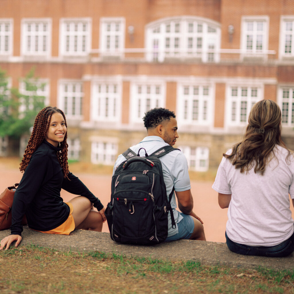 Three people sitting in front of a red brick-layered building with their backs to the camera. One person is turned towards the camera, smiling.