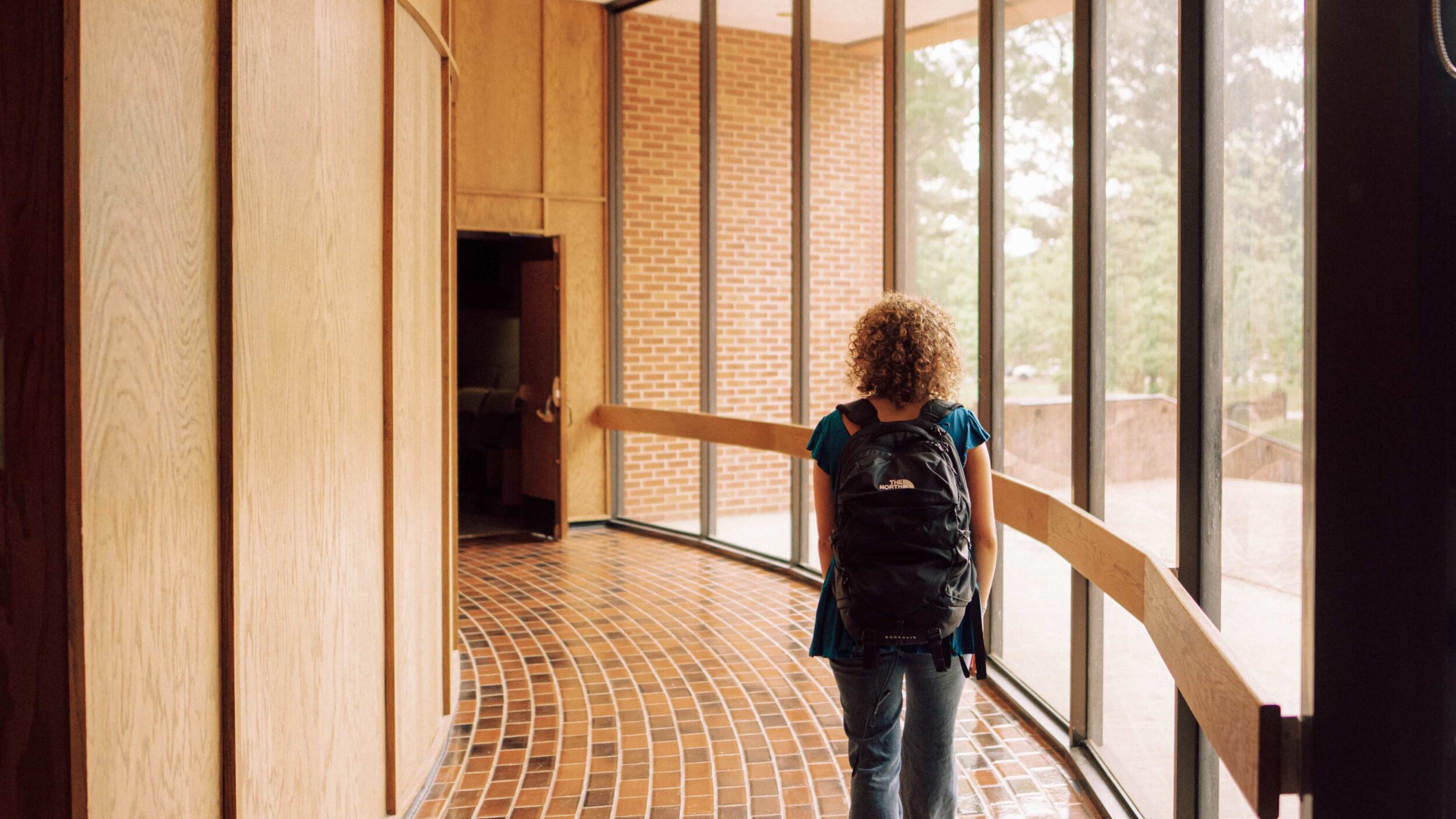 One person walking down a windowed hallway indoors.
