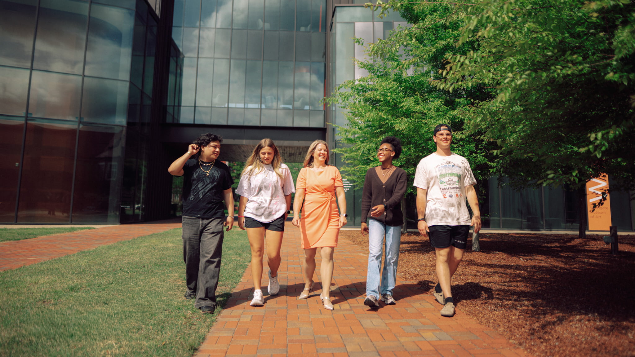 Five people walk outside in front of a glass-paneled building.