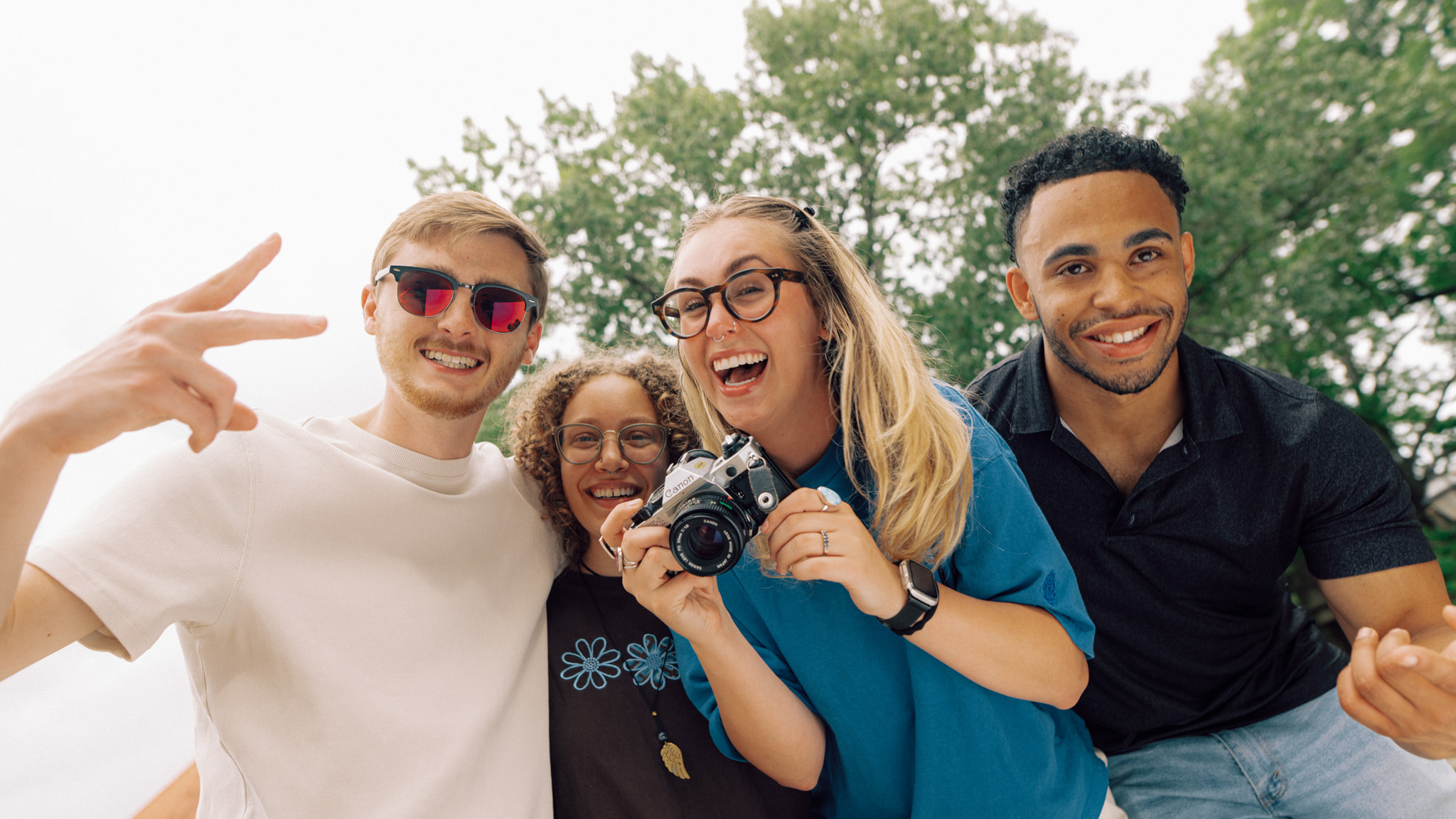 Four smiling people standing beside each other outside. One person is holding a camera.
