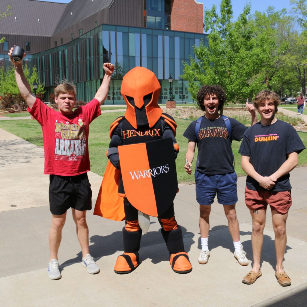 Mascot Altus stands with three students outside on a sunny day. One student has both hands in the air, and another student holds a thumbs-up.