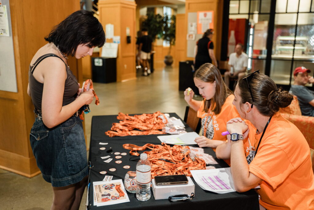 Two people sit at a table that has Hendrix lanyards and stickers laid on it. One person stands in front of them looking at the table.