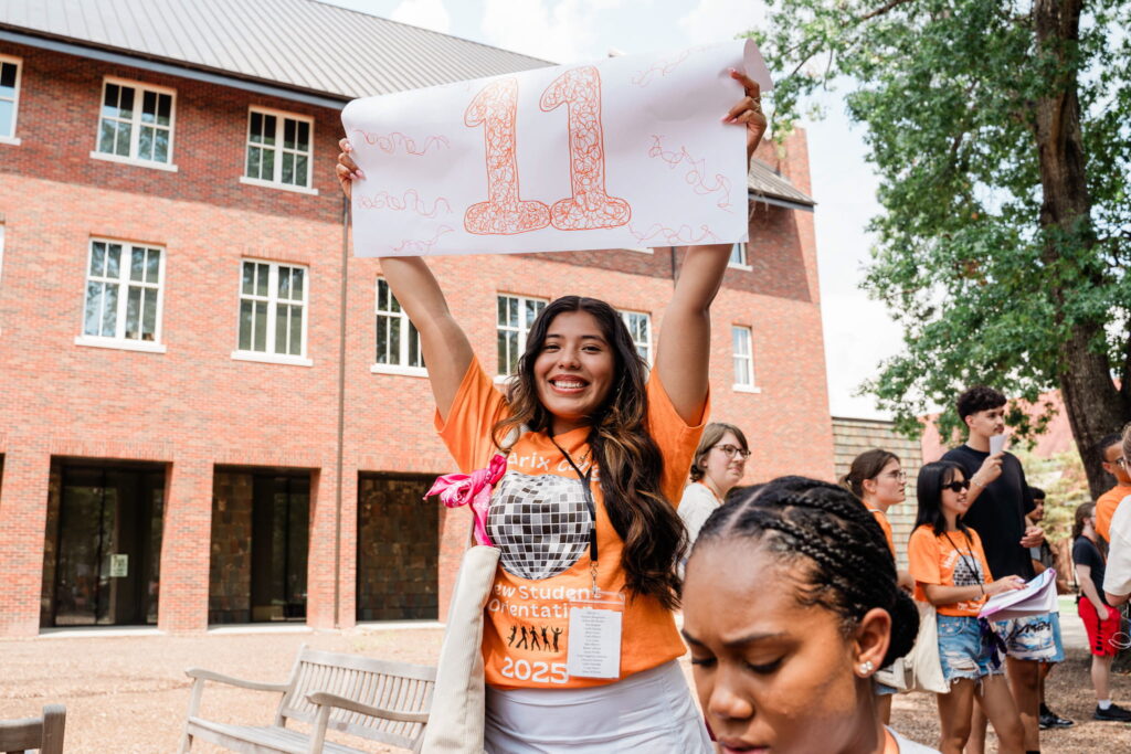 One person smiling while standing outside on a sunny day in front of a red brick building. The person is holding up a sign with the number eleven.