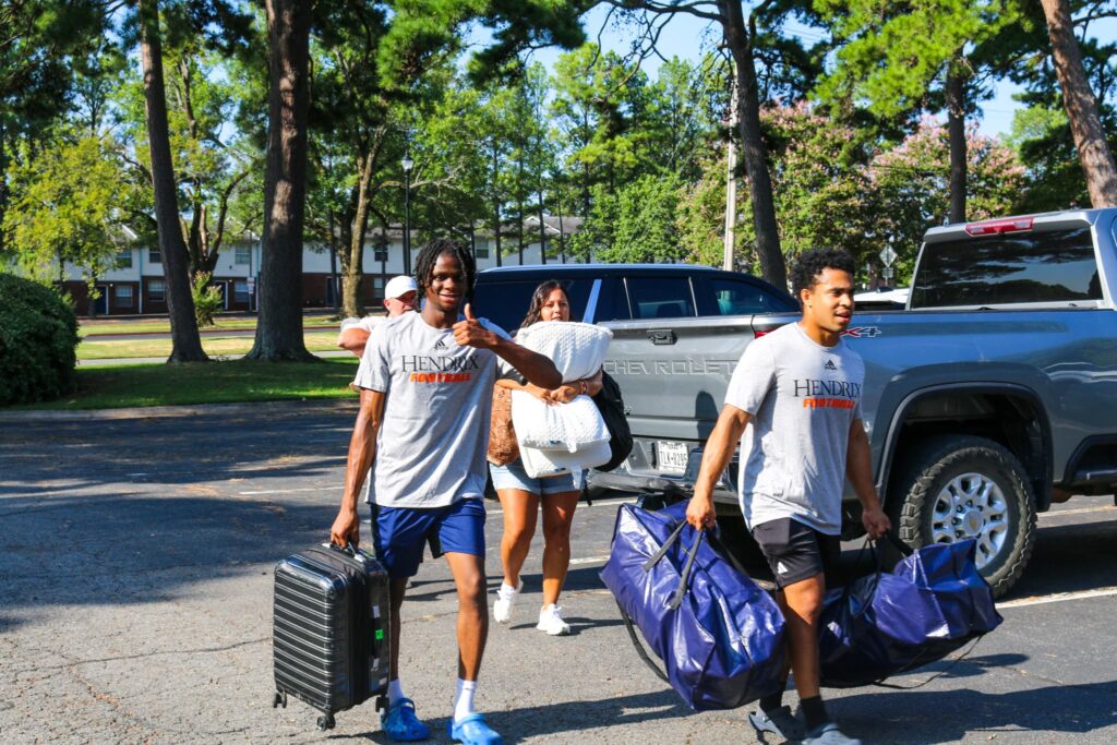 Four people carrying bedding, bags, and suitcases walk in front of two vehicles outside.