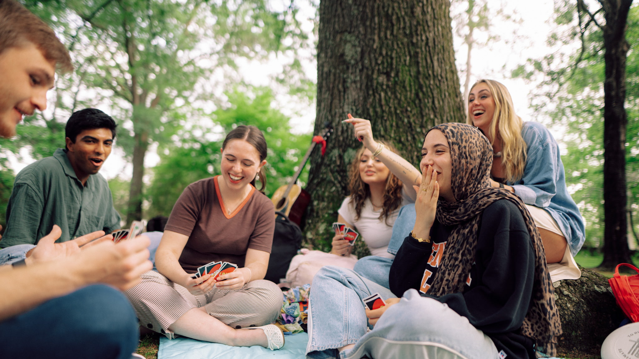 Six students sit around a tree outside playing a card game.
