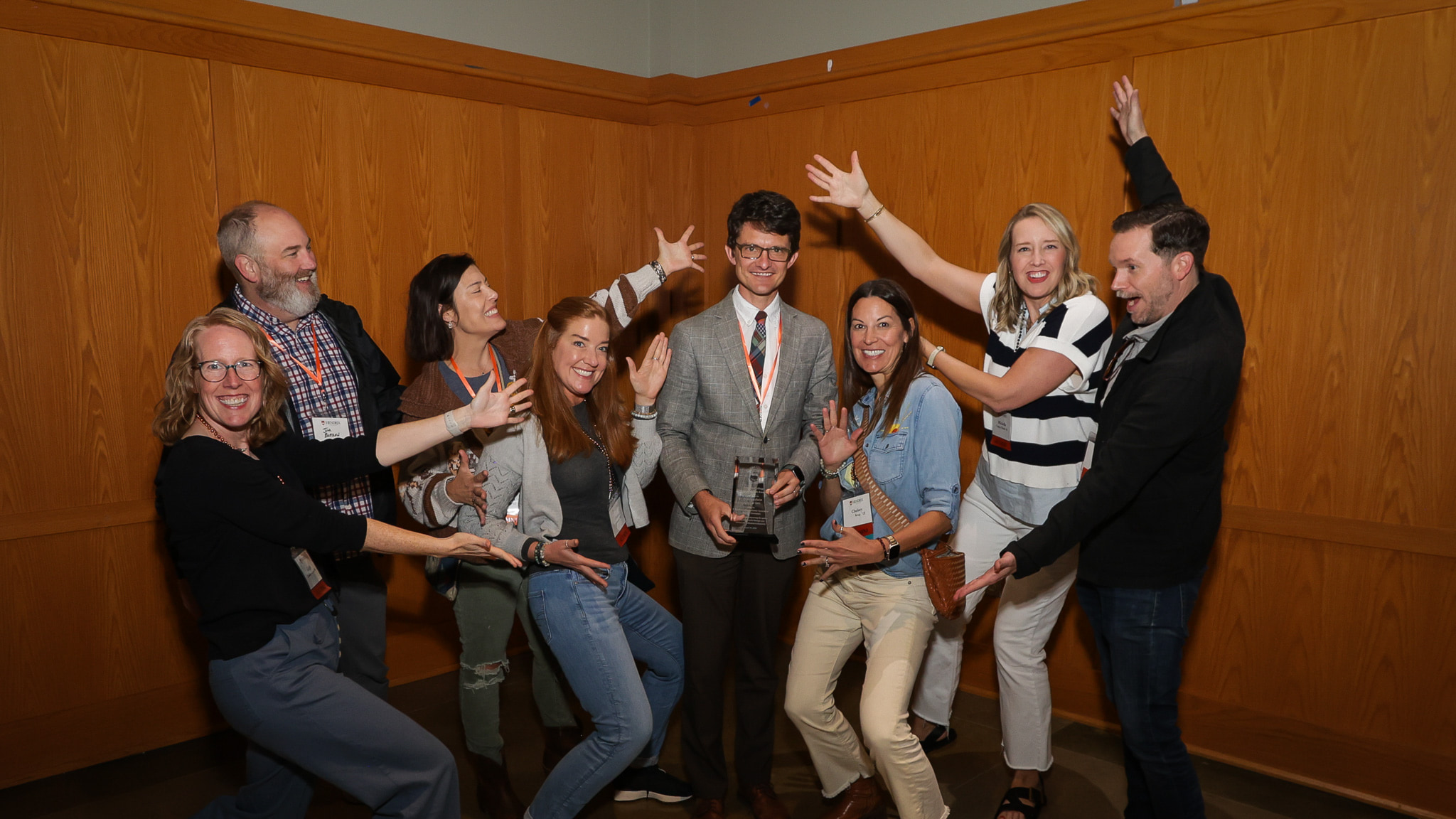 Eight people stand next to each other indoors, smiling with their arms spread wide. One person in the middle holds a glass award.