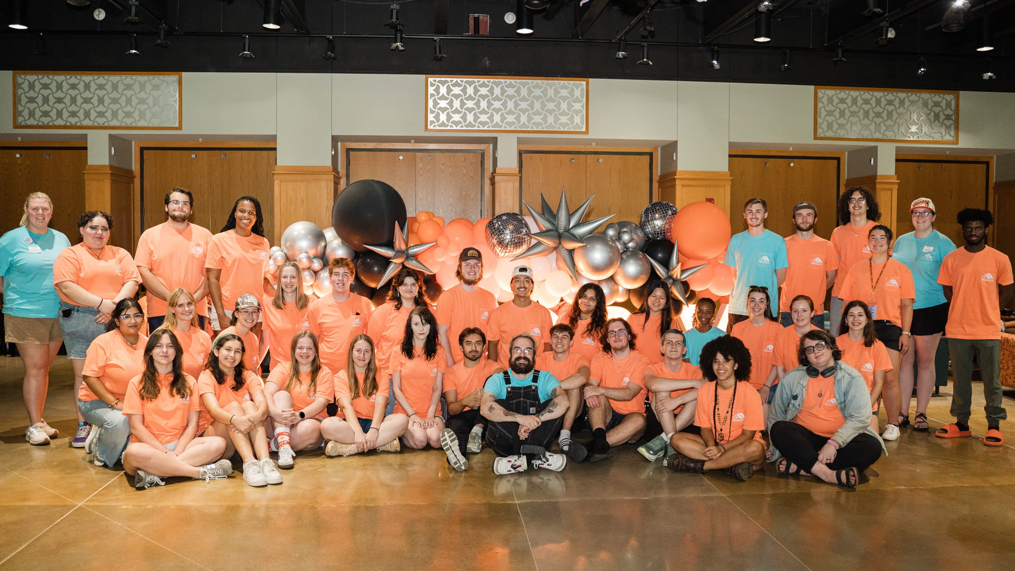 More than 20 people are all wearing orange and blue shirts in a well-lit indoor room. All people are either standing or sitting next to each other.