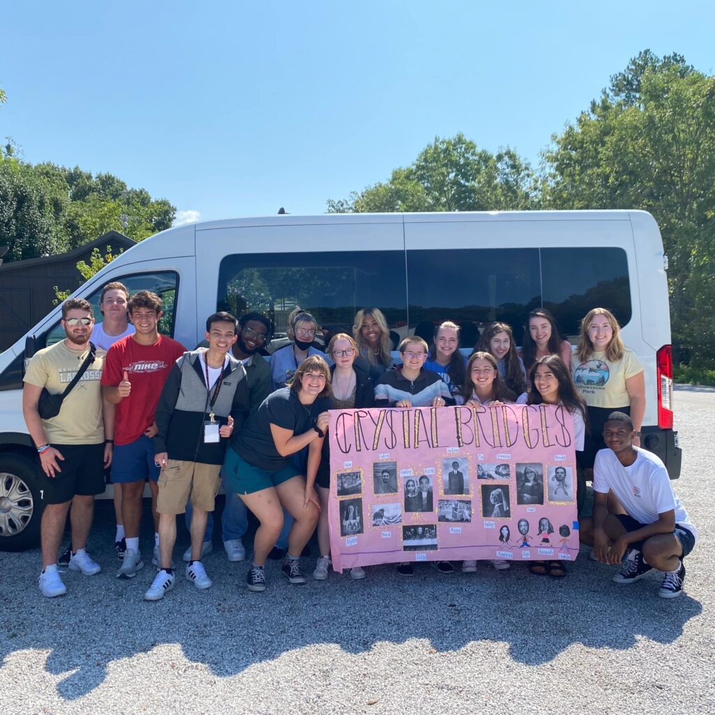 17 students stand holding a pink banner in front of a white passenger van.