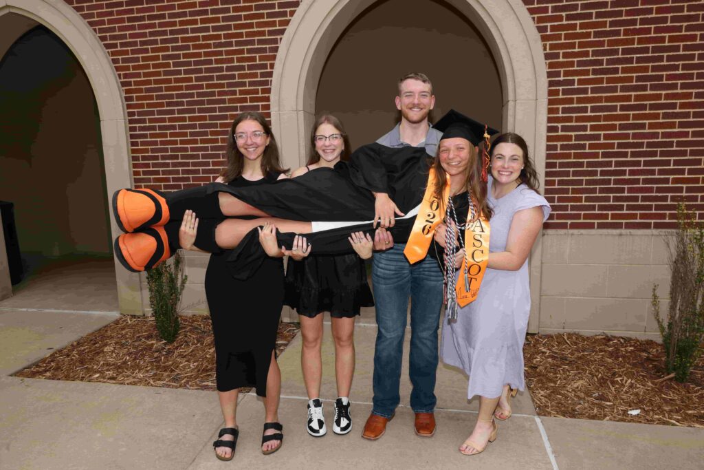 Four students horizontally holding a fifth student who is wearing graduation regalia.