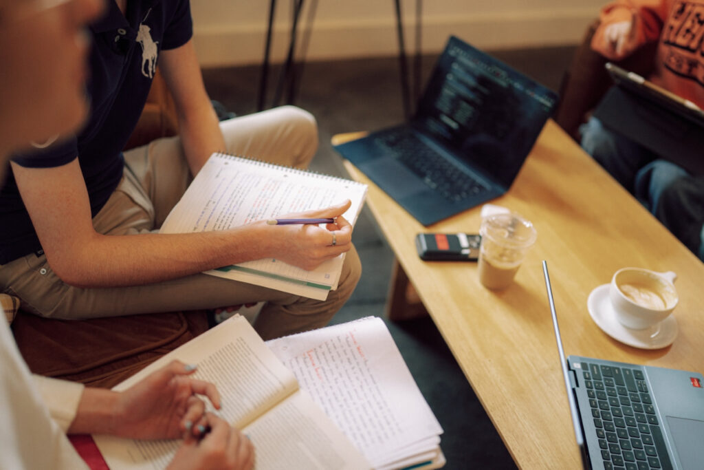 Two students studying at a table with notebooks, electronic devices, and coffee.