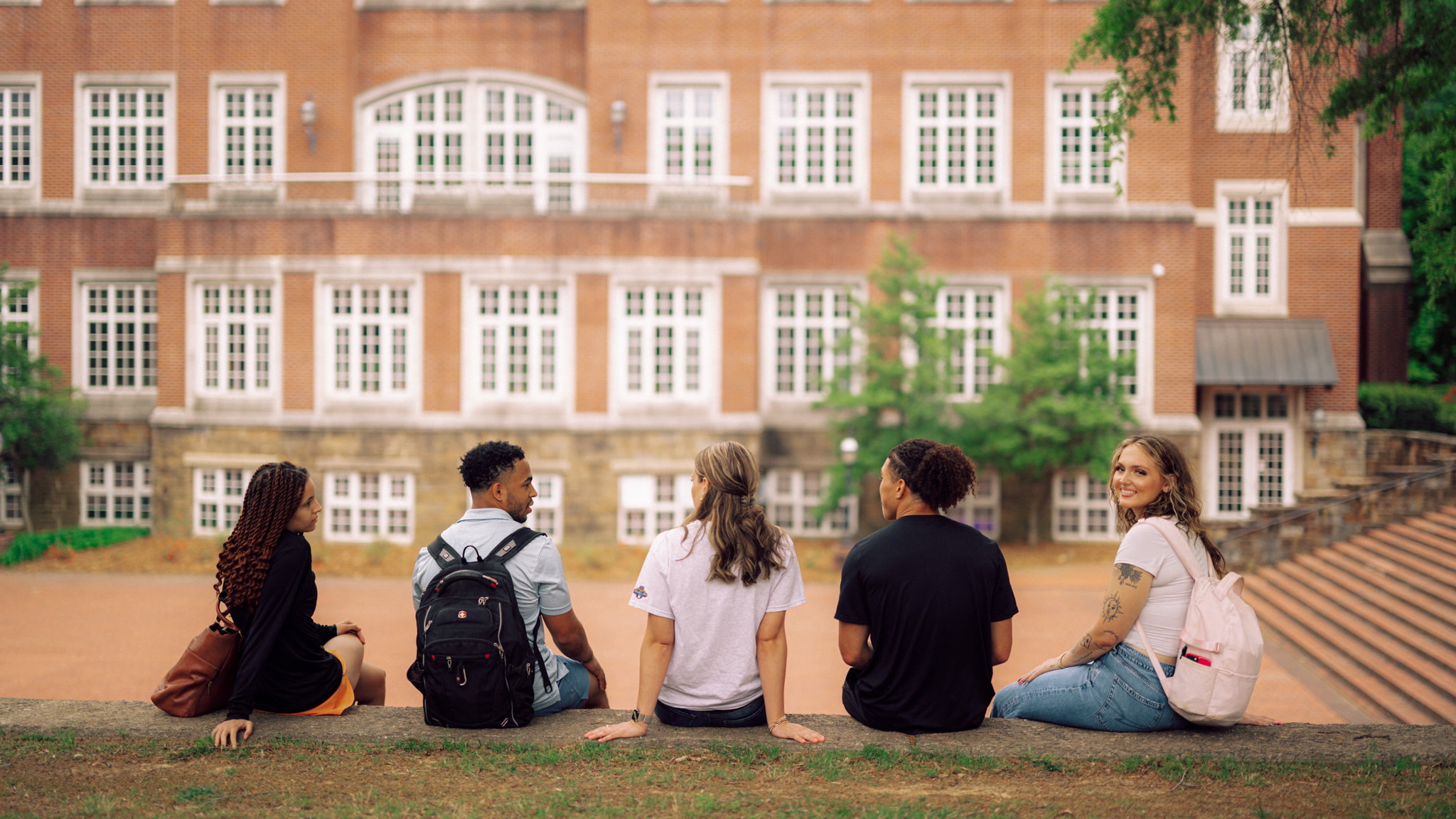Five people sit on a ledge outside with a red brick building across from them.
