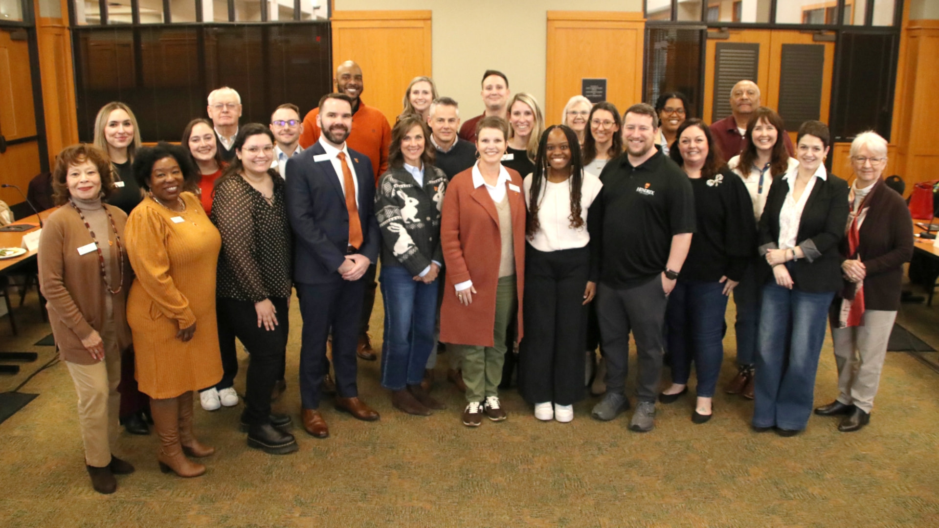Twenty five people standing next to each other smiling in a well-lit indoor room.
