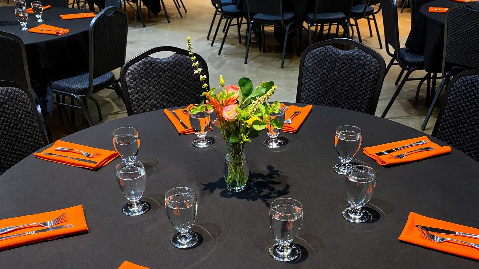 One black circular table, a vase of flowers in the middle, 4 black chairs, drinking glasses throughout the table with silverware and napkins nearby.