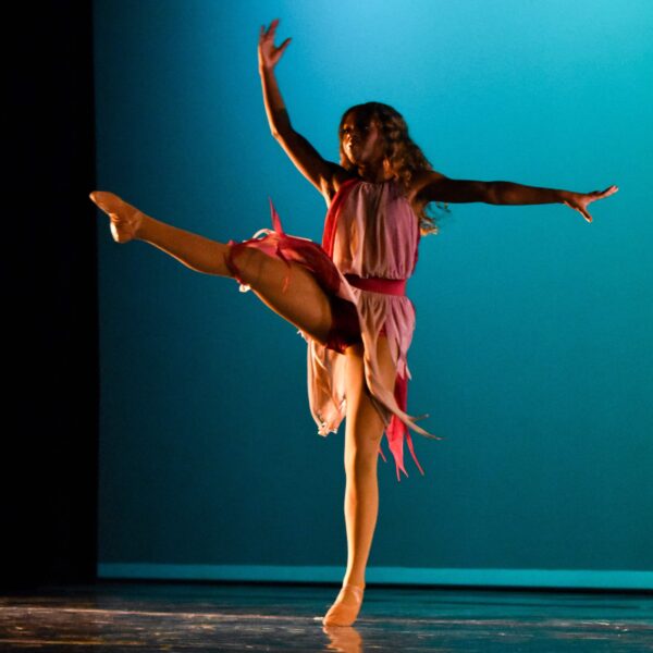 A dancer balances on one leg with the other extended, performing on stage in a flowing pink costume against a blue backdrop.