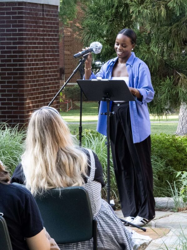 A smiling student adjusts a microphone on a stand while standing at a podium outside in front of an audience.