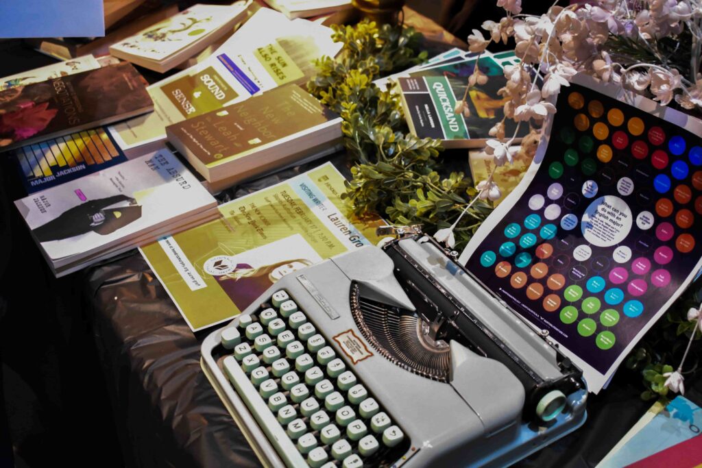 A table with black table cloth covered in a variety of books and a vintage typewriter with a colorful flyer.