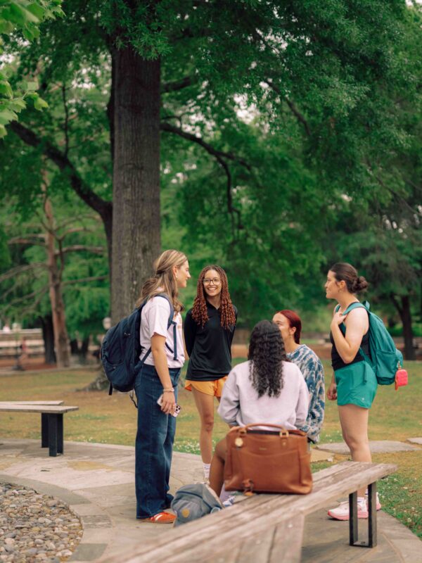Five students outdoors on campus sitting and standing around benches talking.