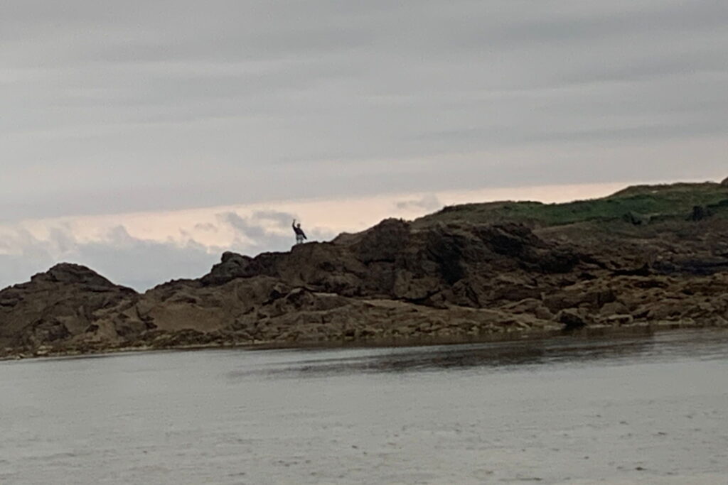 A person in the distance waving while standing on a rock formation of land surrounded by water.