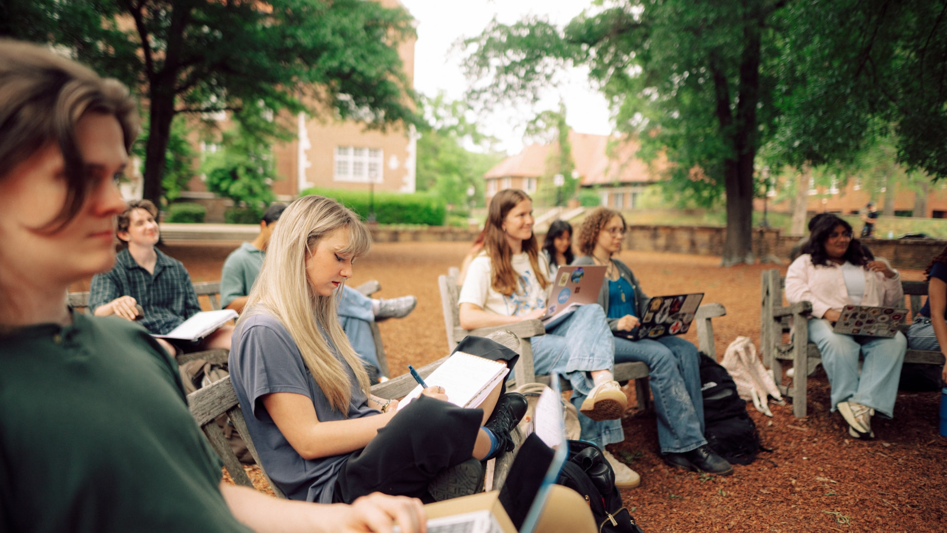 Ten students sit on benches in an outdoor classroom at Hendrix College, taking notes on a sunny afternoon.