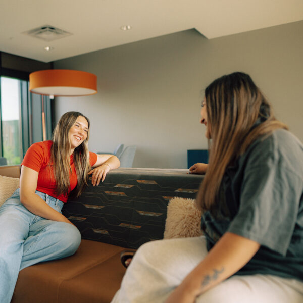 Two Hendrix students sit on a couch in a well-lit room, chatting with one another.