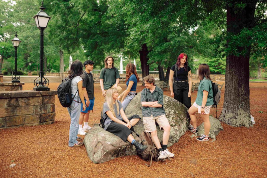 A group of students gather outside under trees near a fountain.